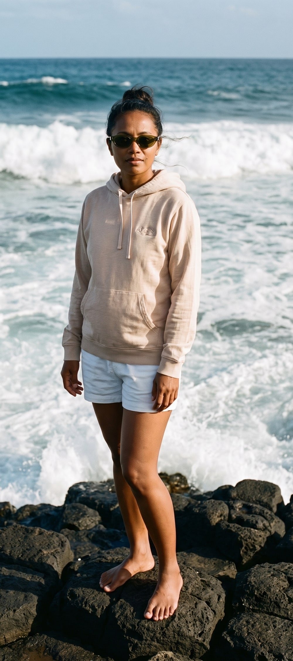 A woman on the basalt rocks at Gris Gris in the olive Alizé sunglasses and oat Riviera Hoodie
