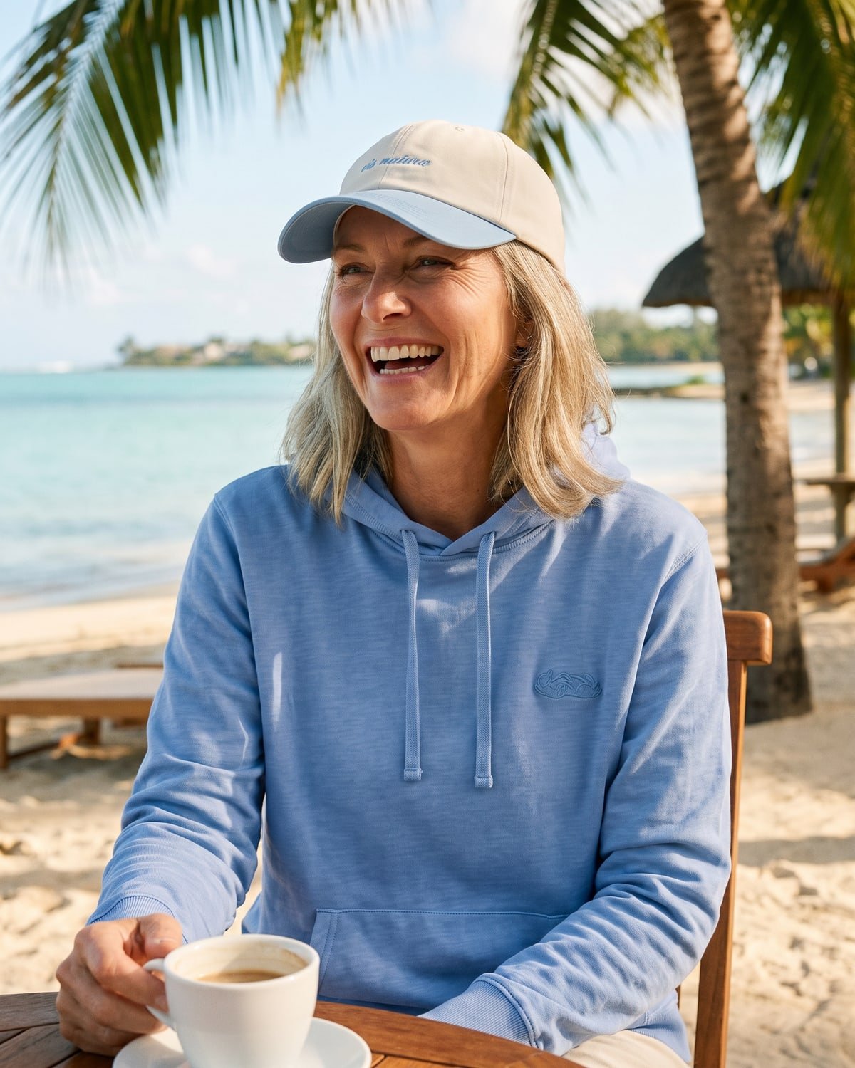 A woman laughing at a café table on the sand at Trou d’Eau Douce in the ecru and sky Marram Cap and sky Riviera Hoodie